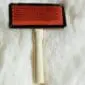 Sheepskin-Rug-Brush-01-1.jpg Sheepskin rug brush with a light wood handle and a rectangular head featuring red metal pins.