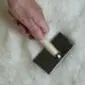 Sheepskin-Rug-Brush-04-1.jpg A hand holding a sheepskin rug brush with a wooden handle and metal bristles against a white wool surface.