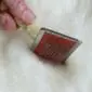 Sheepskin-Rug-Brush-05-1.jpg A hand holding a wooden sheepskin rug brush with metal pins against white wool.