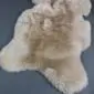 Natural beige sheepskin rug with thick, textured wool and an irregular organic shape on a dark grey background.
